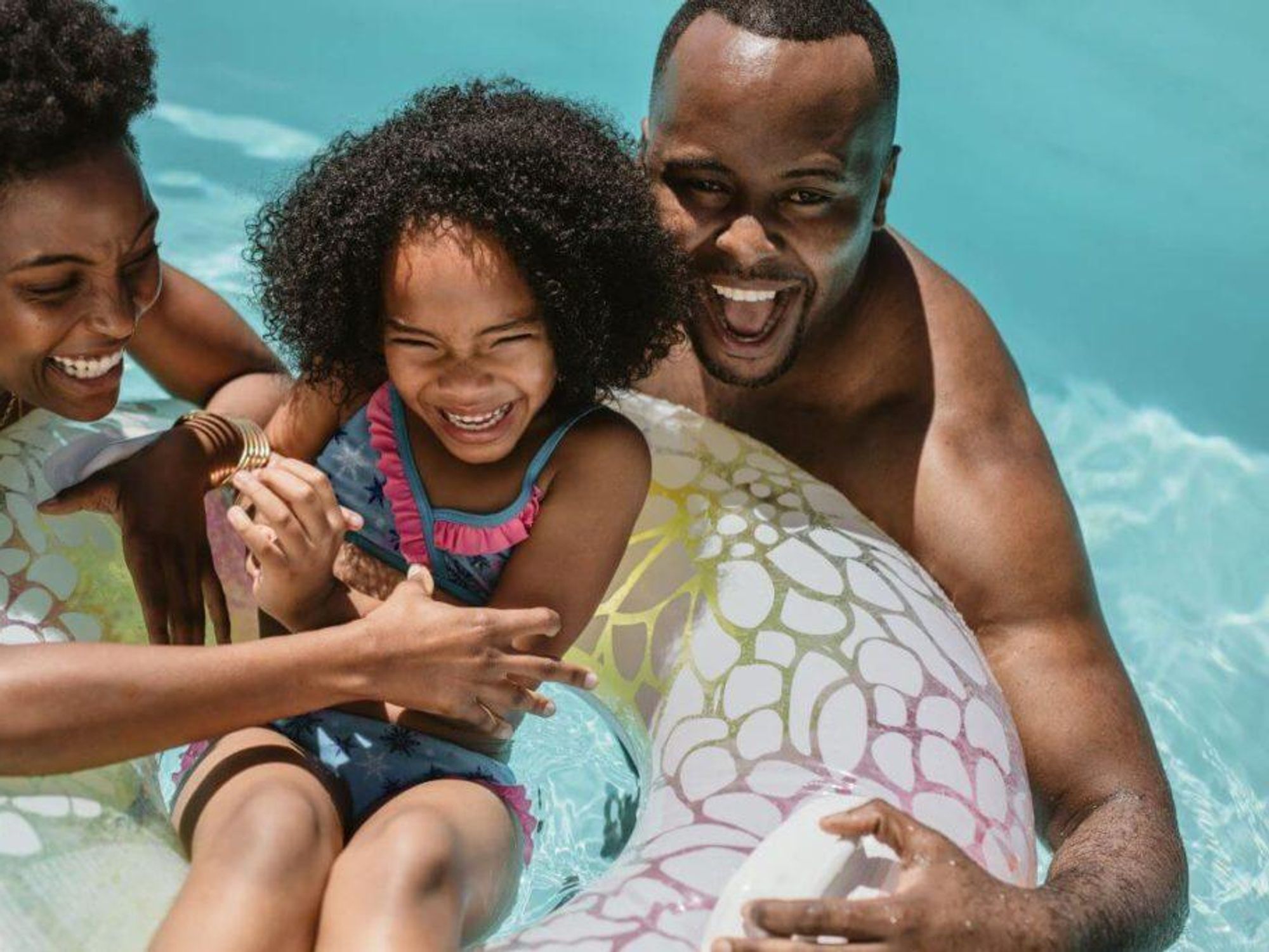 Two parents laugh while holding their child in a floating donut in a pool.